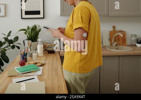 Femme avec le diabète à l'aide de smartphone dans la cuisine avec l'accent sur le dispositif CGM sur le bras Banque D'Images