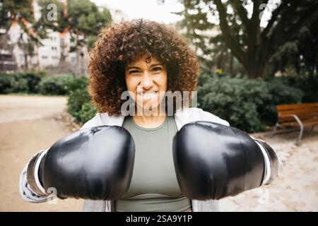 Portrait d'une jeune femme aux cheveux bouclés portant des gants de boxe s'entraînant dans un parc en plein air, exprimant détermination et force. Banque D'Images