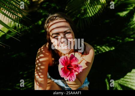 Femme souriante tenant une fleur rose tout en étant assise sur un champ d'herbe. Ombre de feuilles sur son visage. Banque D'Images