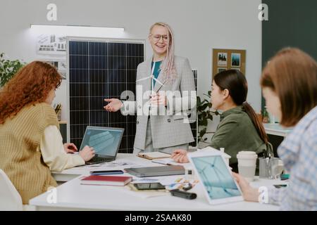 Femme d'affaires souriante donnant une présentation dans un cadre de bureau moderne avec des collègues engagés et discutant à table. La présentation comprend l'affichage de panneau solaire et divers appareils technologiques Banque D'Images