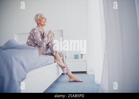 Femme âgée profitant d'une matinée paisible en pyjama tout en étant assise sur le lit dans une chambre lumineuse Banque D'Images