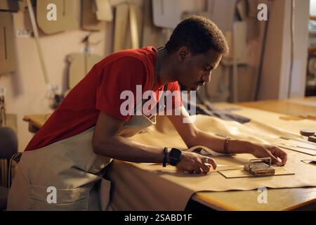 Artisan travaillant méticuleusement sur des pièces en cuir avec une attention focalisée. Homme portant un tablier tout en coupant et façonnant le cuir sur une table en bois recouverte de matériaux Banque D'Images