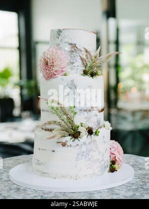 Un gâteau à trois niveaux magnifiquement conçu présente une finition blanche lisse, décoré de fleurs fraîches et de verdure, dans un lieu chic et moderne rempli de wi Banque D'Images