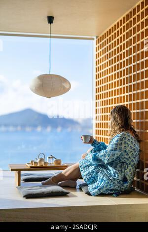Une femme est assise dans un appartement serein, dégustant du thé vert, avec une vue imprenable sur l'océan et les montagnes lointaines à travers une grande fenêtre. Banque D'Images