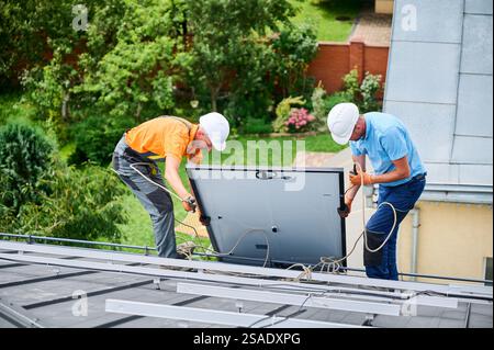 Hommes ouvriers installant un système de panneau solaire sur le toit de la maison. Électriciens en casques soulevant le module solaire photovoltaïque avec l'aide de cordes à l'extérieur. Concept d'énergie alternative et renouvelable. Banque D'Images