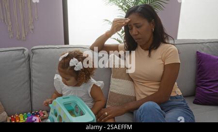 Mère assise stressée sur un canapé dans le salon avec fille jouant à proximité représentant la vie de famille à l'intérieur. Banque D'Images