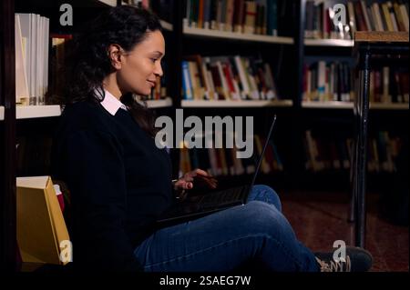 Une femme est assise sur le sol d'une bibliothèque, travaillant sur un ordinateur portable entouré d'étagères. Le cadre met l'accent sur l'apprentissage, la concentration tranquille et le U. Banque D'Images