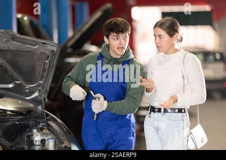 Mécanicien conseille jeune femme cliente sur la réparation sous le capot de la voiture dans la station-service de voiture Banque D'Images