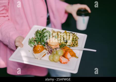 Une personne tient une assiette remplie de poulet grillé, de légumes frais, de pâtes et de café. Le repas coloré suggère une variété de saveurs et c sain Banque D'Images
