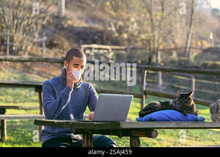 Le jeune homme aime une tasse de café tout en travaillant sur son ordinateur portable à une table de pique-nique dans un cadre rural, avec ses chats dormant paisiblement à côté de lui Banque D'Images