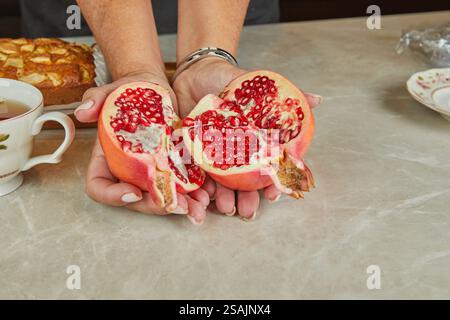 Une personne tient deux moitiés de grenade fraîchement coupées, révélant des graines rouges vibrantes, sur fond de cuisine. Banque D'Images