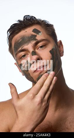 Jeune homme avec des cheveux mouillés et bruns, faisant des procédures faciales, appliquant un masque d'argile nourrissant sur la peau sur fond blanc. Banque D'Images