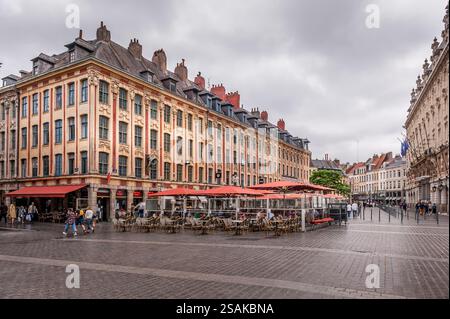 Vue sur une place historique de Lille, France, avec une architecture traditionnelle de style flamand, des cafés avec terrasses, et des gens marchant sous un ciel nuageux. Banque D'Images