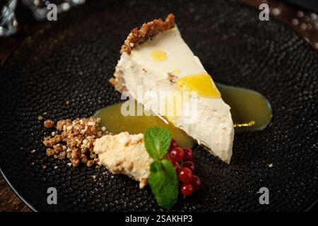 Tranche de cheesecake crémeux avec une croûte de craquelins graham, garnie d'un filet de sauce citron. Garni de feuilles de menthe fraîche, de groseilles rouges et d'un crumble Banque D'Images