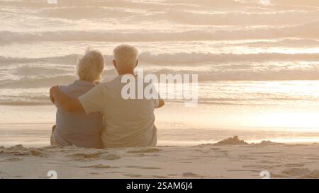 Vue arrière d'un couple de aînés amoureux assis sur le sable sur la plage vacances regardant le coucher du soleil sur l'océan Banque D'Images
