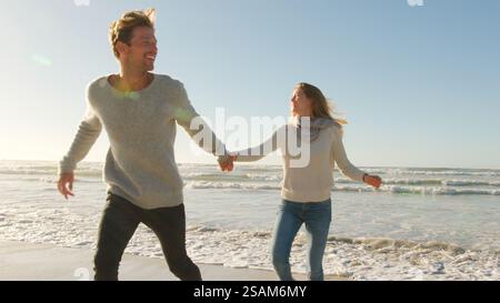 Couple tenant la main sur le rivage de Winter Beach s'enfuyant de briser les vagues Banque D'Images
