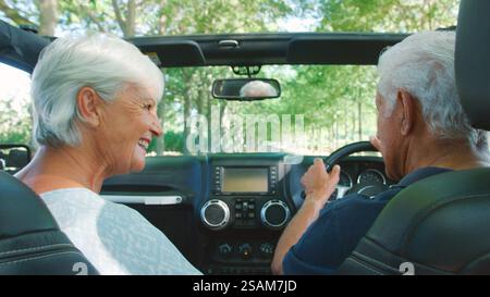 Vue arrière d'un couple sénior sur la route en voiture décapotable conduisant le long de la route bordée d'arbres Banque D'Images