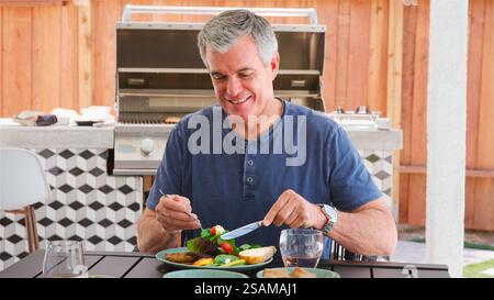 Portrait d'un homme senior à l'extérieur à la maison assis à table manger barbecue repas avec salade Banque D'Images