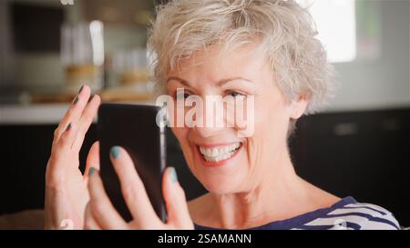 Femme senior sur canapé à la maison en utilisant le téléphone mobile navigation en ligne streaming Show ou vérification des messages Banque D'Images