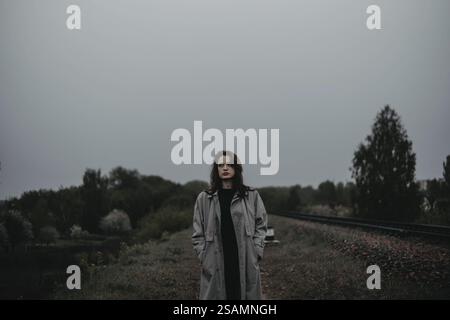 Une femme vêtue d'un manteau se tient à l'extérieur près des voies ferrées sous un ciel couvert, créant une atmosphère contemplative Banque D'Images
