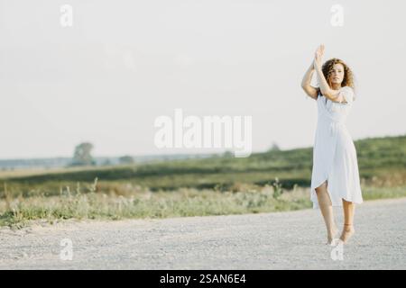 Une femme dans une robe blanche danse gracieusement sur une route de campagne sous la lumière du soleil Banque D'Images