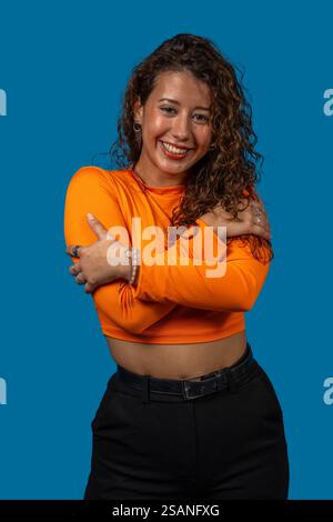 Jeune femme latino-américaine souriante avec les cheveux bouclés se serrant dans ses bras, portant une chemise orange et un pantalon noir, sur un fond bleu Banque D'Images