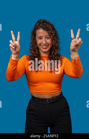 Portrait studio d'une jeune femme souriante faisant signe de paix avec les deux mains sur un fond bleu Banque D'Images