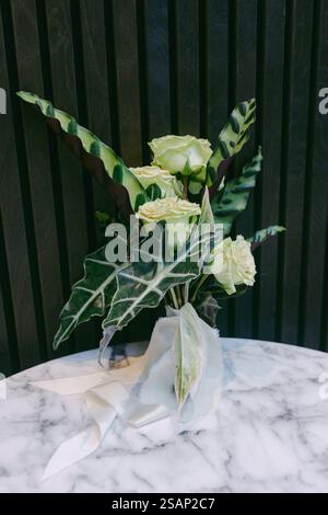 Un bouquet magnifiquement arrangé avec des roses vert clair et des feuilles tropicales vibrantes se trouve sur une table en marbre. Les textures contrastées créent un Banque D'Images