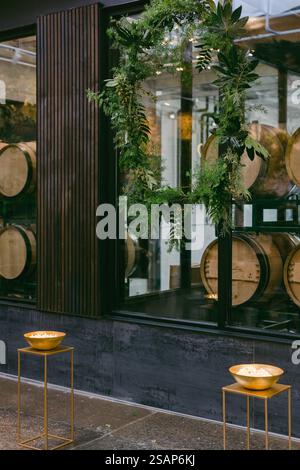 Une grande couronne verte orne l'entrée d'un magasin de vin, entourée de tonneaux en bois brillants empilés dans la fenêtre. Deux bols élégants reposent sur des s dorés Banque D'Images