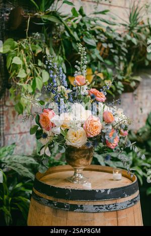 Un bel arrangement floral avec des roses de couleur douce et des fleurs bleues vibrantes est placé sur un tonneau en bois, entouré d'une végétation luxuriante. Le se Banque D'Images