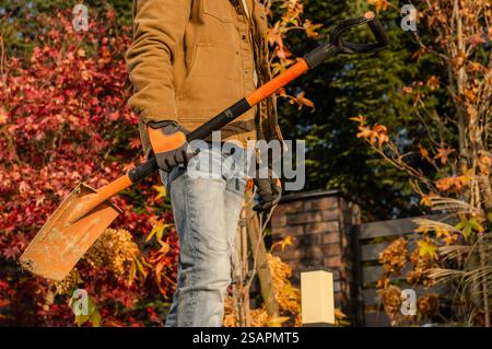 Un jardinier tient une pelle en automne, entouré d'un feuillage rouge et orange vif, se préparant à travailler dans la cour sous un ciel clair. Banque D'Images