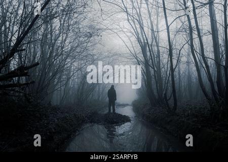 Figure solitaire effrayante, debout dans une forêt effrayante par un jour d'hiver brumeux, sa silhouette reflétée dans l'eau. Banque D'Images