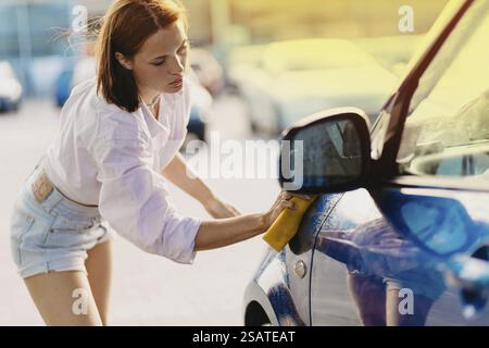 Femme frottant le côté de la voiture avec une éponge jaune par une journée ensoleillée Banque D'Images