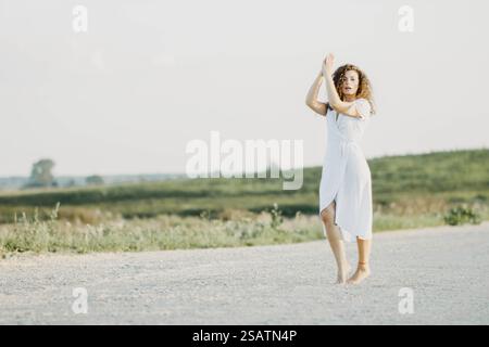 Une femme dans une robe blanche danse pieds nus sur une route de campagne paisible Banque D'Images