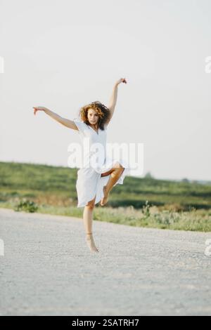 Femme bondissant joyeusement dans une robe blanche sur une route tranquille Banque D'Images