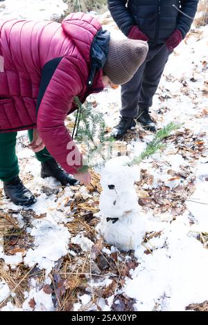 Deux enfants décorent soigneusement un petit bonhomme de neige avec des rameaux à feuilles persistantes dans un cadre forestier hivernal. Le sol est recouvert de neige et de feuilles séchées. Banque D'Images