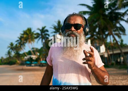 Homme senior sud-asiatique avec la barbe grise aime la musique, danse sur la plage tropicale. Porter des écouteurs, des lunettes de soleil, embrasse un style de vie actif. Homme âgé Banque D'Images
