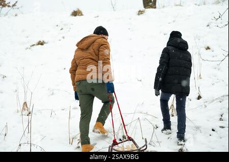 Deux personnes, groupées en tenue d'hiver, marchent en montée à travers un paysage enneigé. Une personne tire un traîneau en bois derrière eux, profitant d'une journée d'hiver outdo Banque D'Images