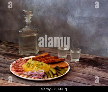 Une carafe en verre et deux verres de vodka froide sur une table en bois, à côté d'une assiette avec des pommes de terre, du saindoux, des concombres, du hareng et du pain de seigle. Gros plan. Banque D'Images