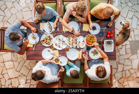 Vue de dessus de la famille joyeuse réunie autour de la table à manger en plein air dégustant un repas méditerranéen avec du poisson frais, des légumes et des salades, des plats vibrants o Banque D'Images