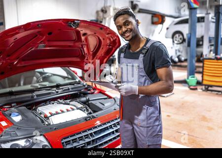 Mécanicien souriant prenant des notes sur le presse-papiers tout en inspectant le moteur d'une voiture rouge avec capot ouvert dans un atelier professionnel Banque D'Images
