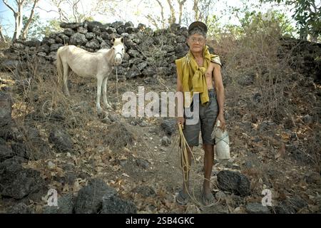 Portrait d'un homme âgé debout, alors qu'il tient une corde pour conduire un poney sur un paysage rocheux dans le village traditionnel de Prailiang dans le Mondu, Kanatang, Sumba est, Nusa Tenggara est, Indonésie. On estime que 55 millions de personnes dans le monde sont directement touchées par les sécheresses chaque année, ce qui en fait le danger le plus grave pour le bétail et les cultures dans presque toutes les régions du monde, selon la Convention des Nations Unies sur la lutte contre la désertification (UNCCD) dans sa publication de mai 2022 intitulée «sécheresse en chiffres 2022 : restauration pour la préparation et la résilience». Banque D'Images