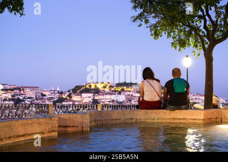 Couple assis pendant l'heure bleue la nuit au belvédère Sao Pedro de Alcantara, surplombant le château de Sao Jorge en arrière-plan. Lisbonne. Banque D'Images
