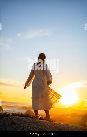 Une femme dans une robe blanche fluide se tient sur une colline rocheuse, regardant un coucher de soleil vibrant. Sa robe attrape la lumière tandis que le soleil illumine l'horizon Banque D'Images