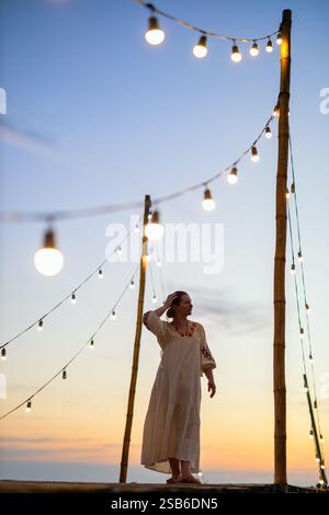 Une femme dans une robe brodée blanche se tient sur une plate-forme sous des lumières décoratives, silhouettée contre un ciel de coucher de soleil vibrant avec des couleurs douces Banque D'Images