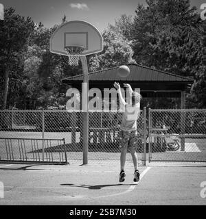 NISSWA, MN - 12 juillet 2024 : terrain de basket-ball de quartier avec un jeune athlète jouant, tirant la balle au panier, dans une photo en noir et blanc. Banque D'Images