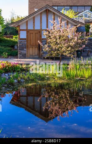 Une maison avec un toit blanc et une porte en bois se trouve devant un étang. L'étang est entouré d'un jardin verdoyant Banque D'Images