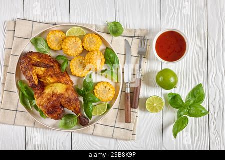 la moitié de poulet rôti avec des pièces d'épis de maïs et des feuilles de basilic frais sur une assiette sur une table en bois blanc avec couverts, citron vert et sauce chili doux, horizonta Banque D'Images