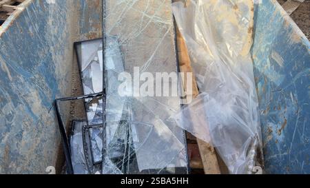 Une benne à ordures bleue contient des vitres brisées dans un cadre noir, accompagnées d'une feuille de plastique et de planches de bois. Le verre présente de vastes fr Banque D'Images