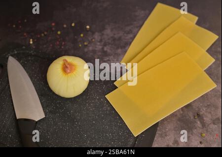 Feuilles de pâtes oignon, couteau et lasagne sur une planche à découper noire de cuisine. Fond noir. Préparation de cuisson des lasagnes maison Banque D'Images